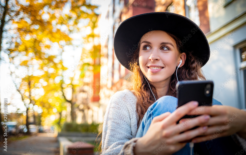 Beautiful stylish woman sitting on street at sunny day and using mobile phone with headphones