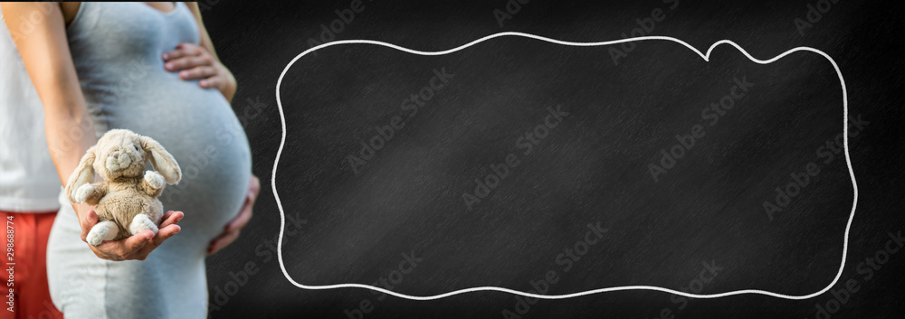 Pregnant woman holds a cuddle toy rabbit in front of her belly, copy space on a blackboard