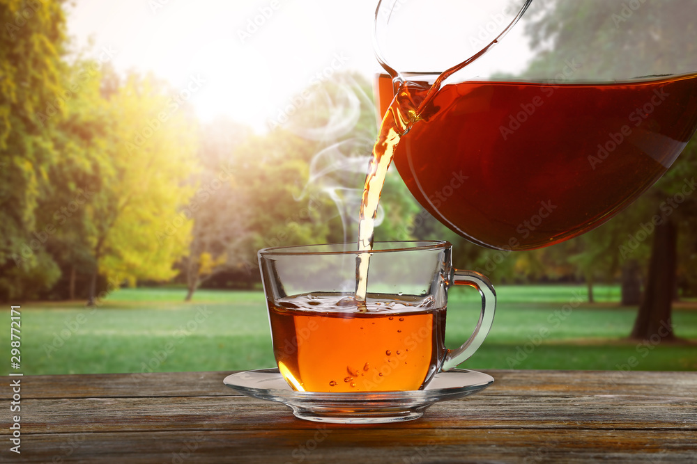 Pouring of tasty hot tea into cup on wooden table in beautiful park