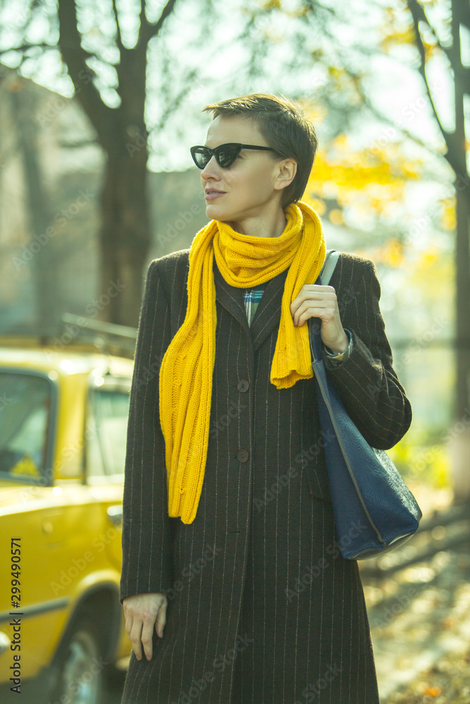 Beautiful autumn woman wearing trendy sunglasses, coat, yellow scarf in the street with yellow car o