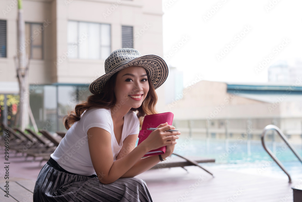 Pretty Asian woman sitting near swimming pool and holding notebook.