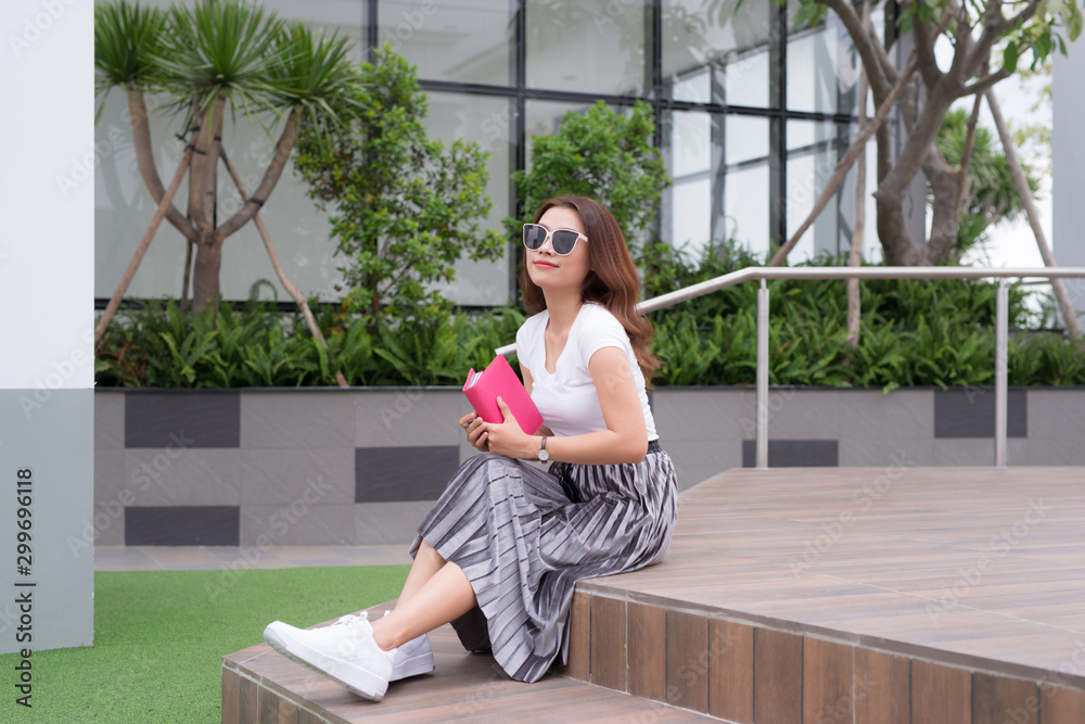 Happy young woman wearing sunglasses sitting on stairs and writing notebook outside.