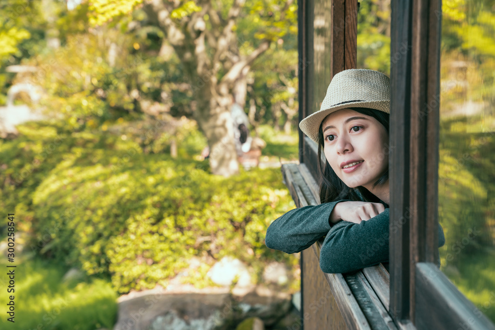 戴着帽子的年轻迷人的女性旅行者靠在窗台上，头朝外呼吸新鲜空气