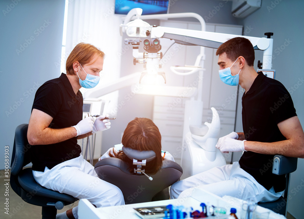 Dentists with protective mask and gloves repair tooth of young female client at ambulant. Oral care.