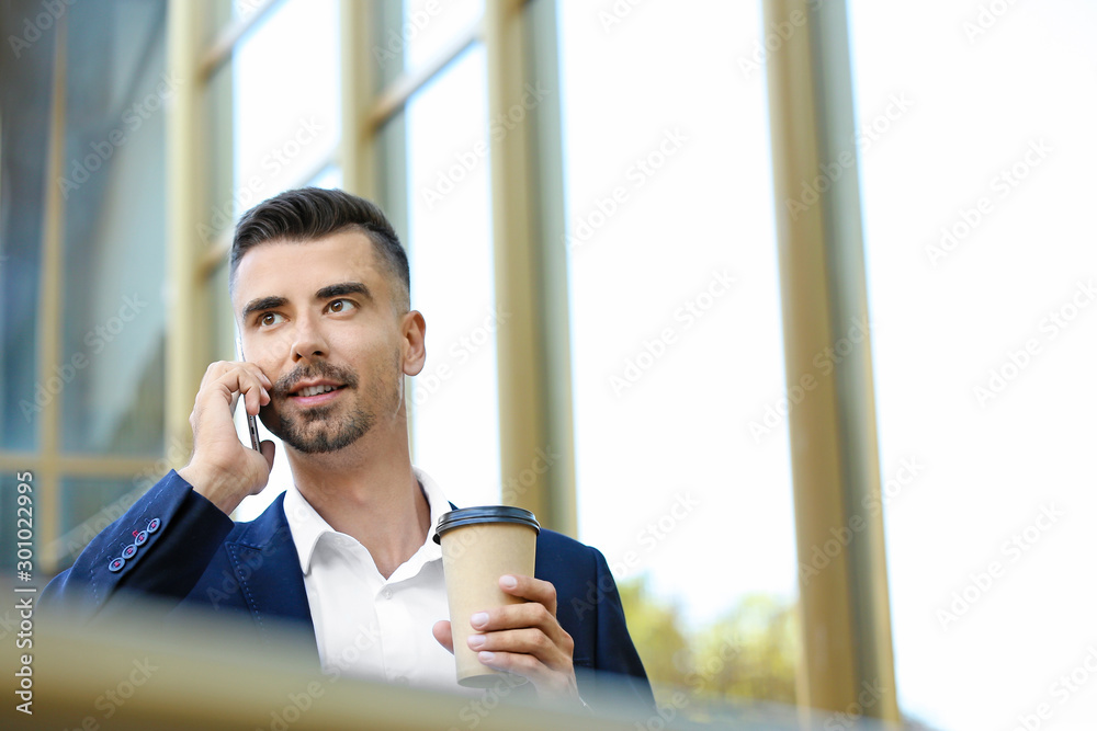 Portrait of handsome businessman with cup of coffee talking by phone outdoors