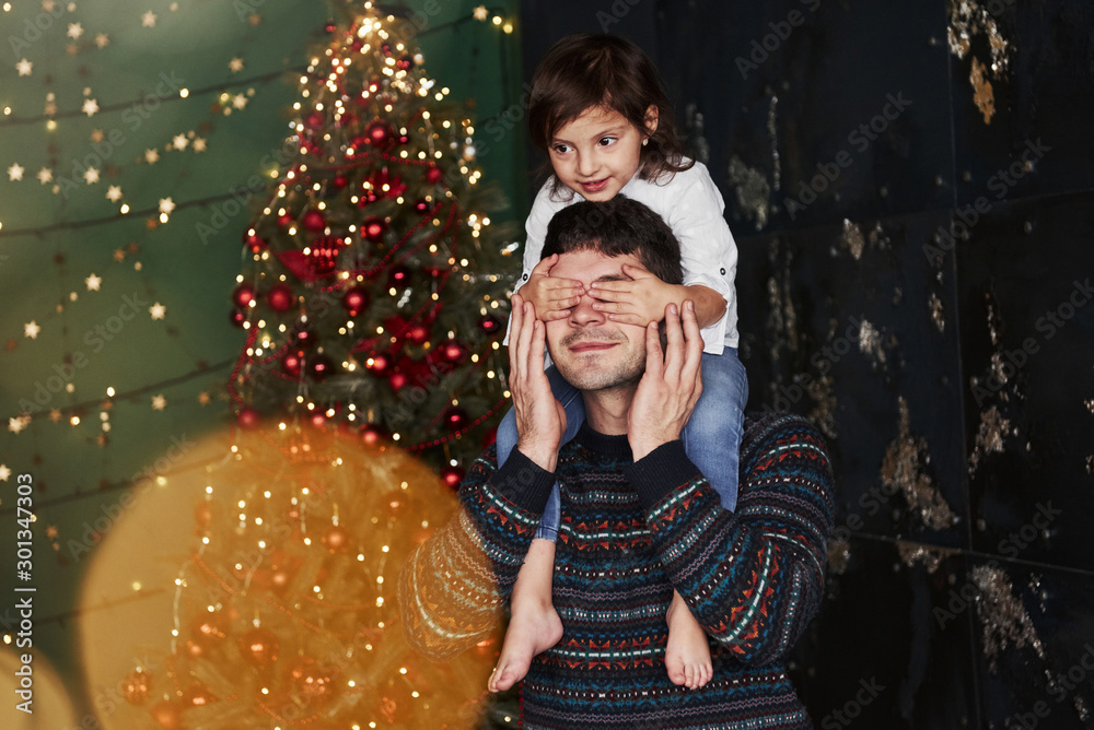 Girl sits on the fathers shoulders and covering his eyes. Decorated Christmas at background