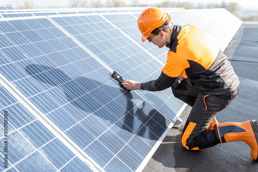 Well-equipped worker in protective orange clothing installing solar panels, measuring the angle of i