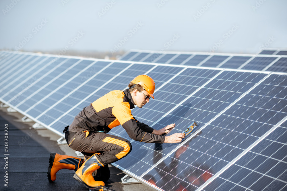 Well-equipped worker in protective orange clothing installing solar panels, measuring the angle of i