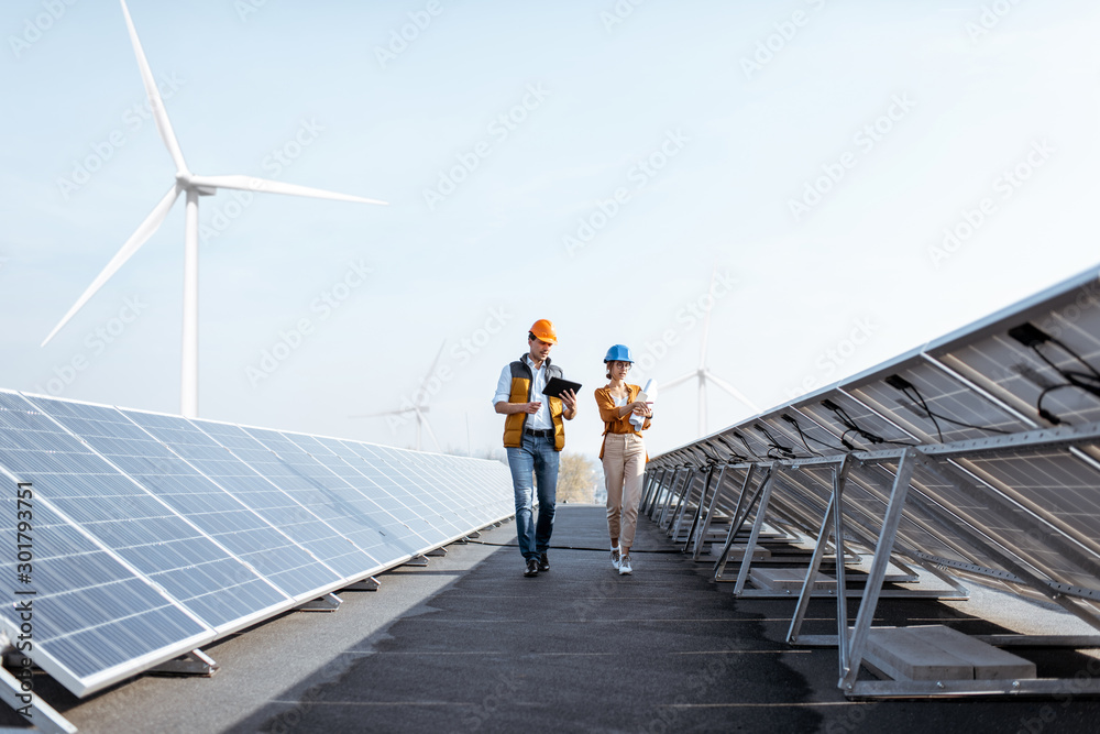 View on the rooftop solar power plant with two engineers walking and examining photovoltaic panels. 