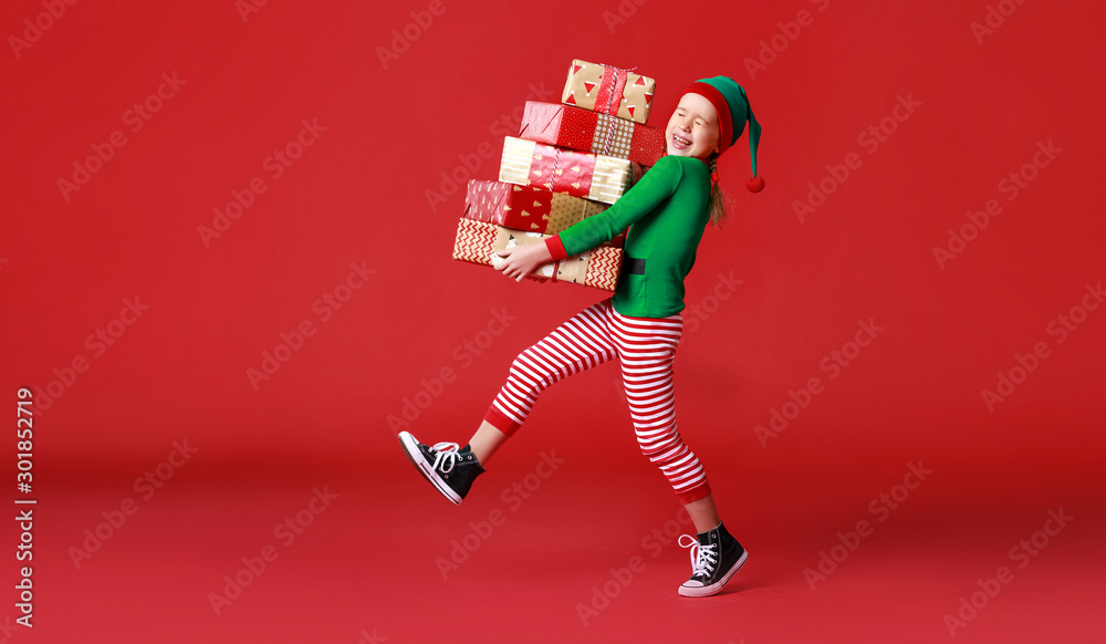 cheerful funny child in Christmas elf costume with gifts on   red background.