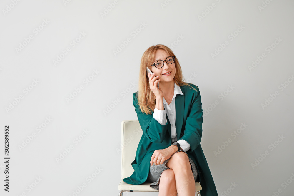 Portrait of stylish mature businesswoman talking by phone while sitting on chair against light backg