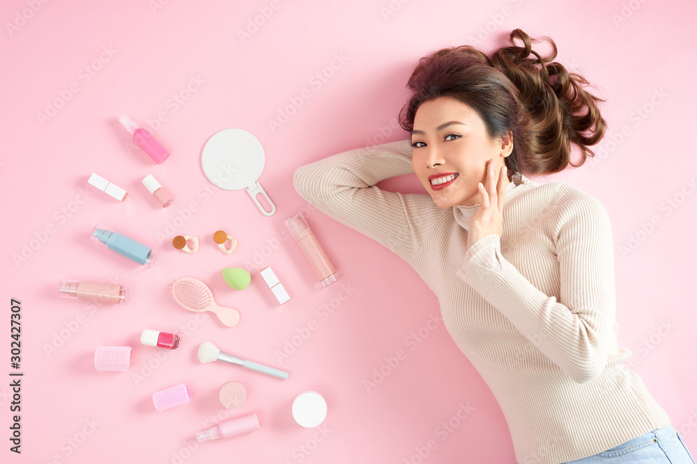 Young Asian girl smiling with makeup cosmetic tool and lying on pink background. Top view