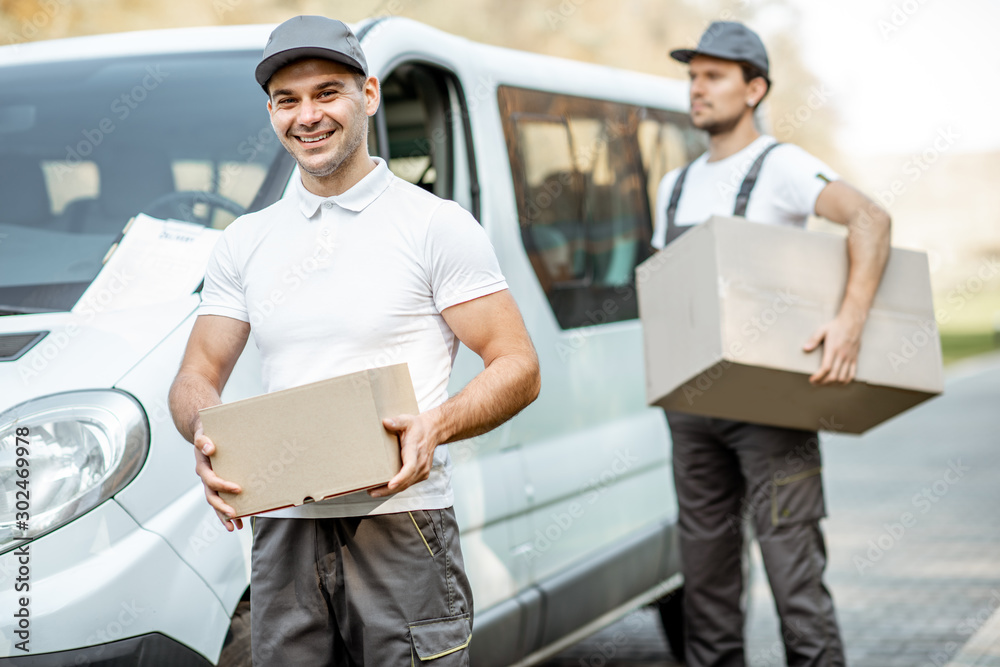 Two cheerful male couriers in uniform delivering goods by cargo van vehicle, unloading cardboard par