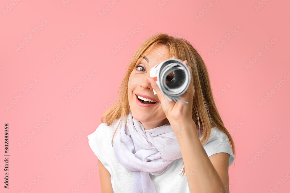 Stylish mature woman looking at viewer through rolled newspaper against color background