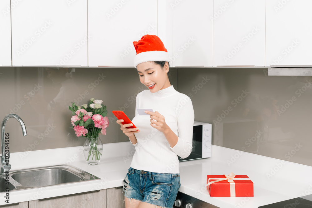 Concept of buying presents on Christmas. Asian woman with Christmas hat at home holding credit card