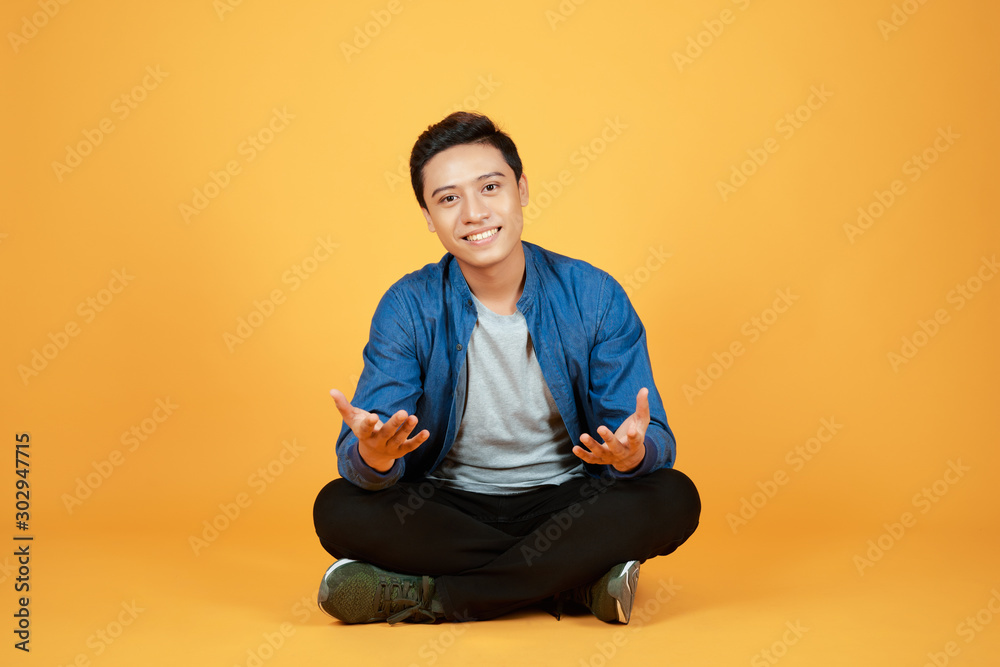 Good looking young Asian man sitting with legs crossed on the floor, isolated on orange background.