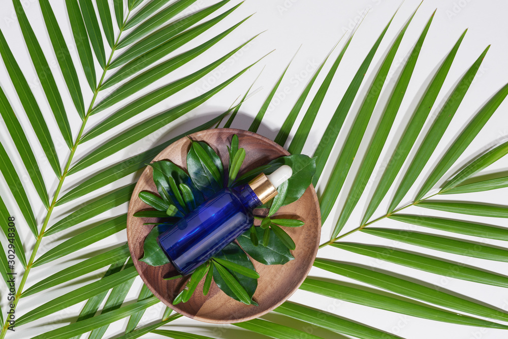 Blue serum bottle on wood plate with green leaves on white background. Spa and beauty concept.