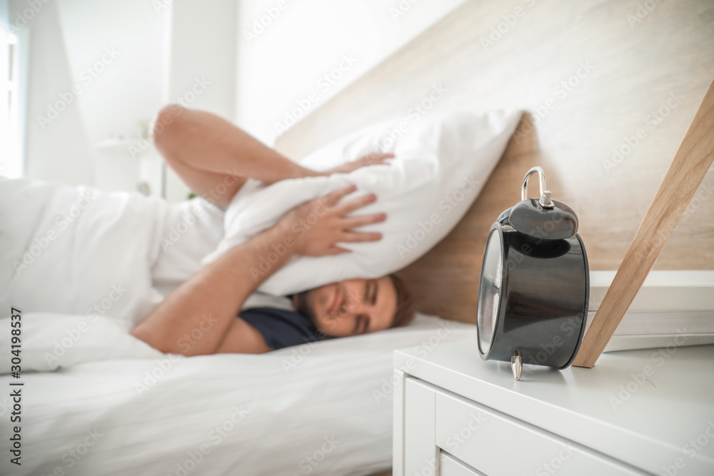 Alarming clock and young man covering head with pillow while trying to sleep in bed