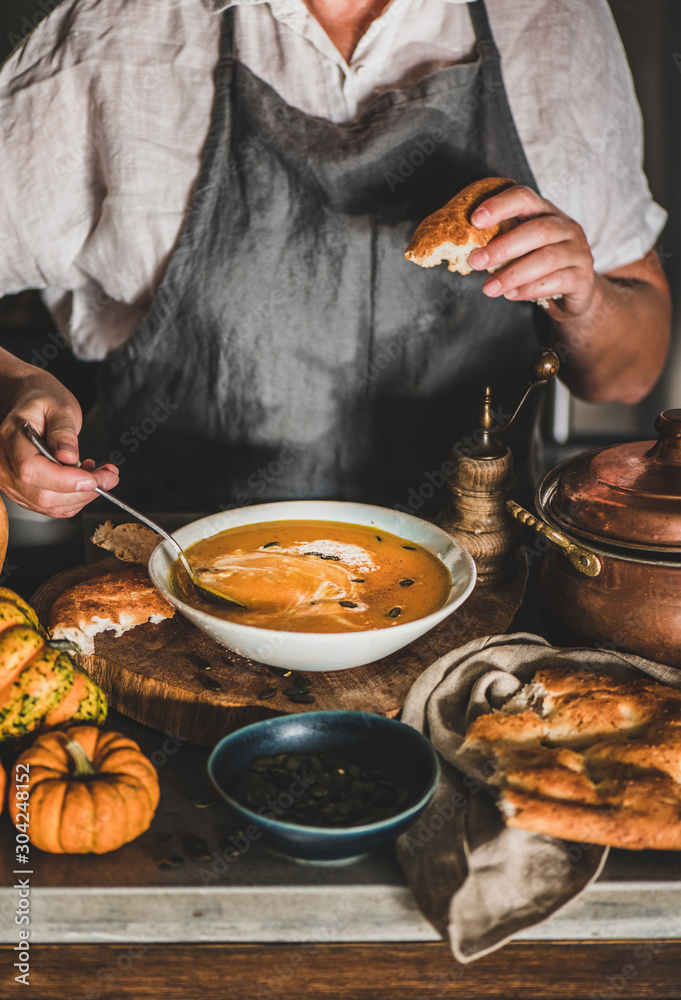 Eldery woman in grey linen apron mixing cream with Autumn seasonal pumpkin cream soup in modern kitc