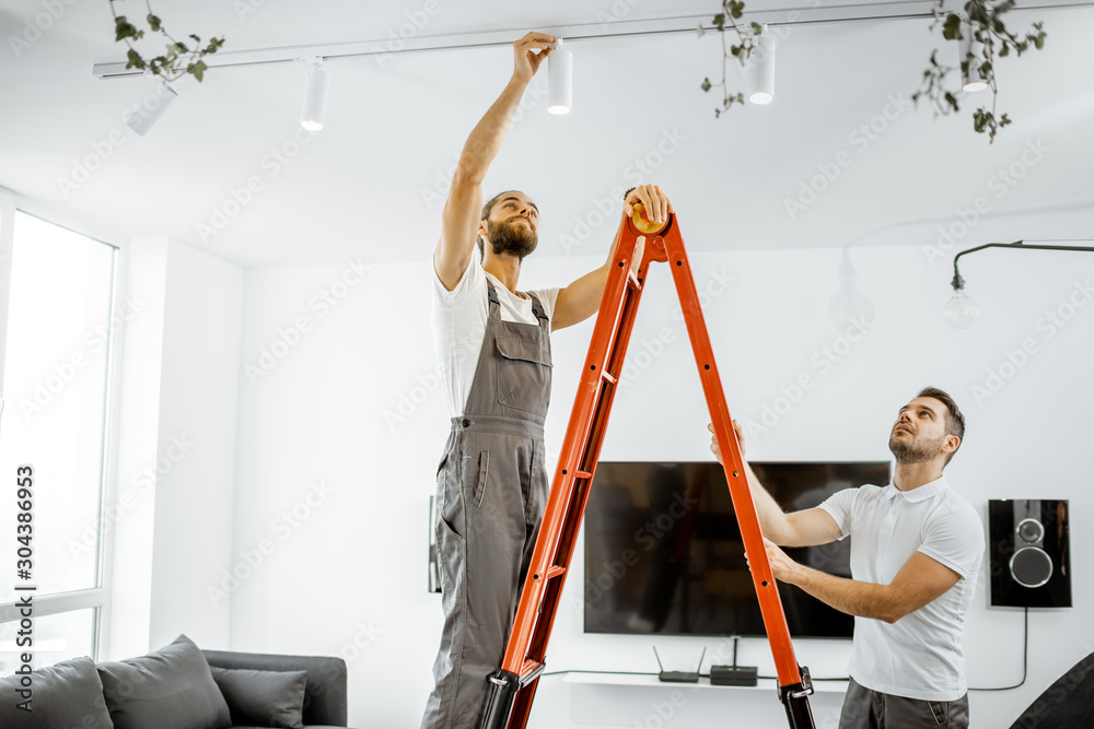 Two repairmen or professional electricians installing light spots, standing on the ladder in the liv