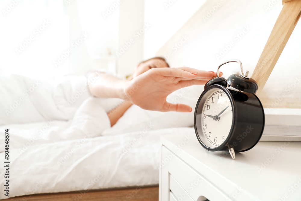 Morning of young woman turning off alarm clock while sleeping in bed