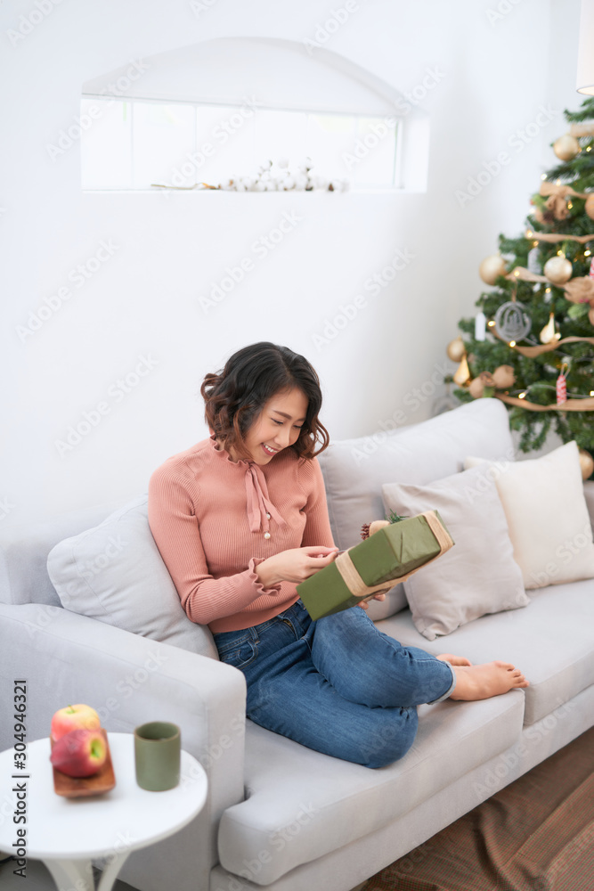 一名年轻女子在圣诞树旁拿着一份礼物。女孩坐在那里微笑着，手里拿着送给圣诞节的礼物