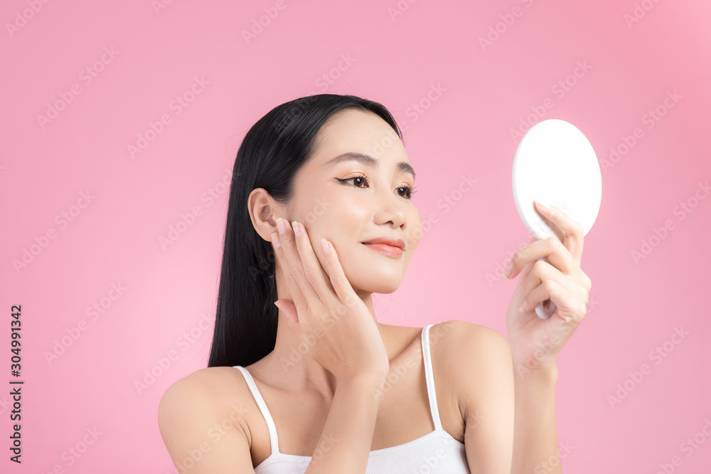 Attractive young Asian woman holding mirror and feeling happy about her skin. Isolated on pink backg