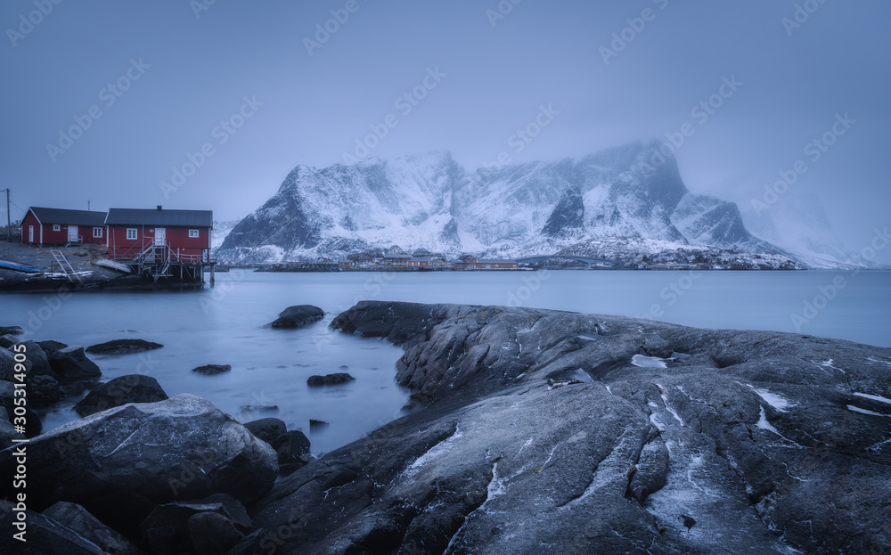 Beach with stones in blurred water, red rorbu and snowy mountains in fog at dusk in winter. Sea coas