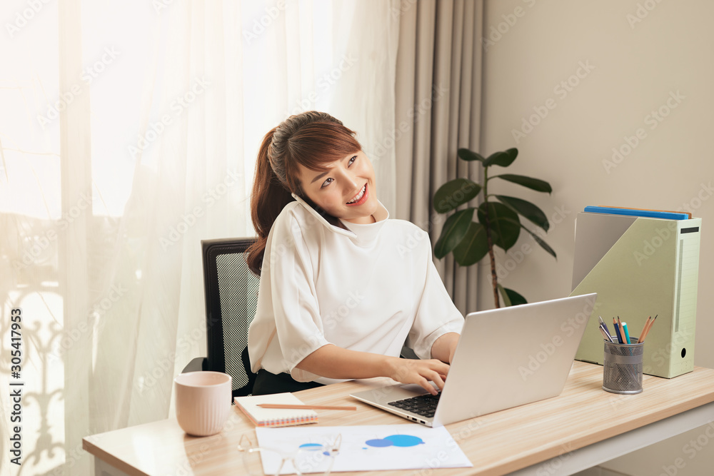 Confident Asian woman talking on her mobile phone and using laptop while sitting at desk and working