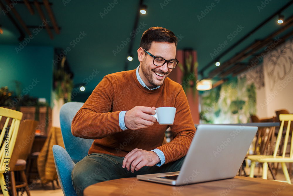 Smiling man drinking coffee and looking at laptop in the cafe, portrait.