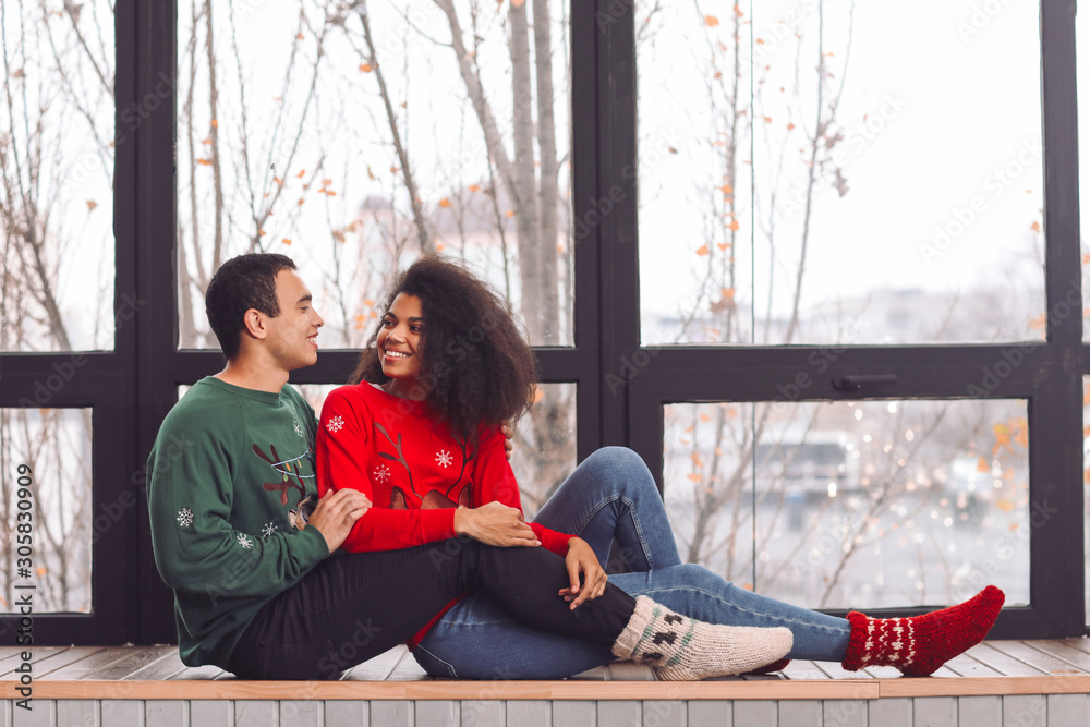 Young African-American couple in Christmas sweaters sitting on window sill