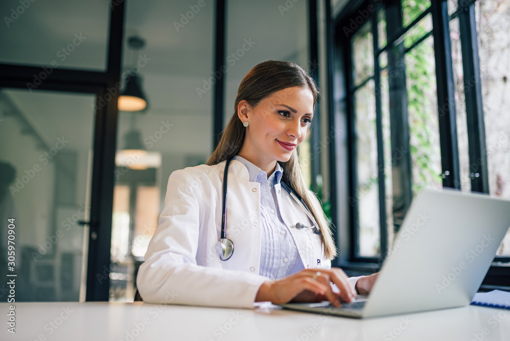 Positive doctor working on laptop in medical office, portrait.