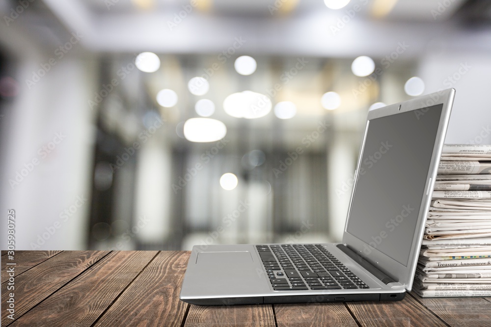 Laptop and newspapers lay on a wooden desk on blurred room background