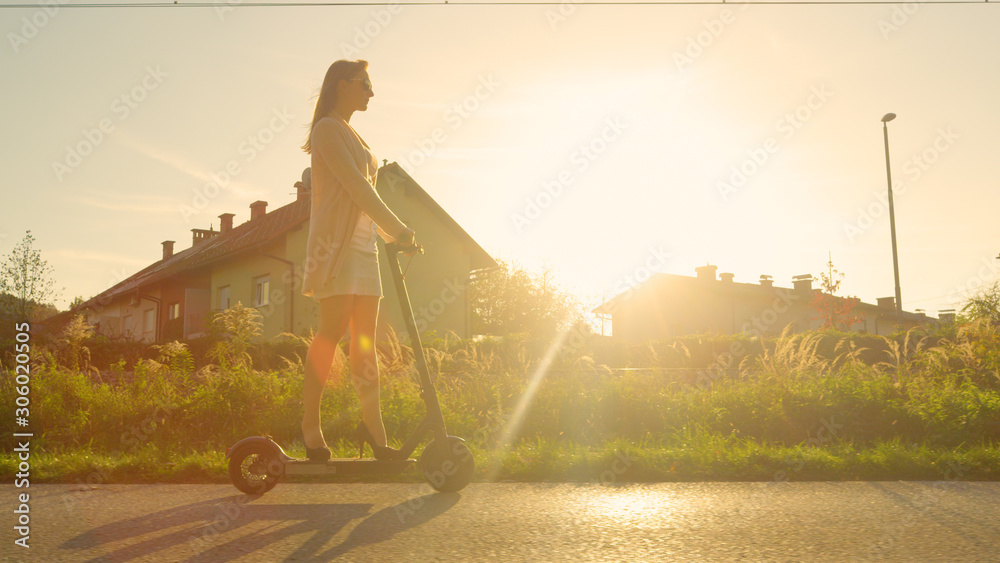 LENS FLARE: Young woman rides an electric scooter past suburban town at sunset.