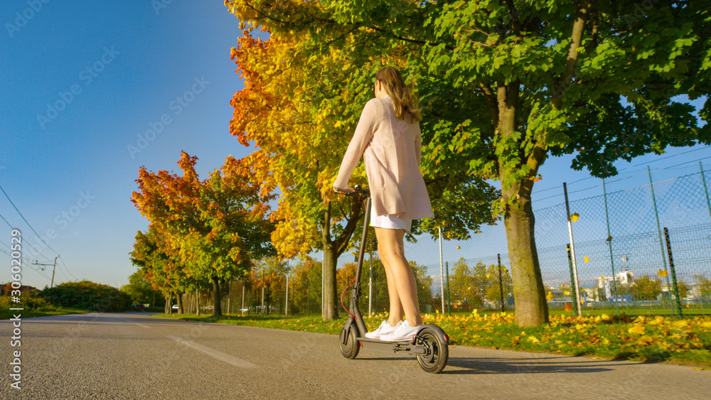 LOW ANGLE: Modern woman riding an electric scooter through the suburbs in autumn