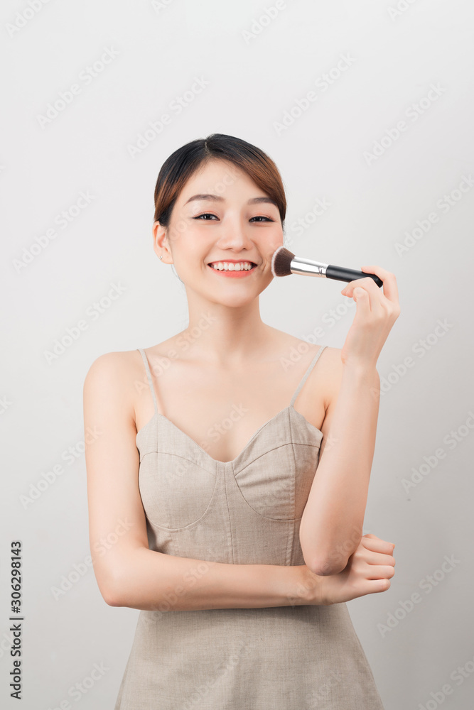 Cheerful woman is doing make up on her face using brush. Beauty routine on white background.