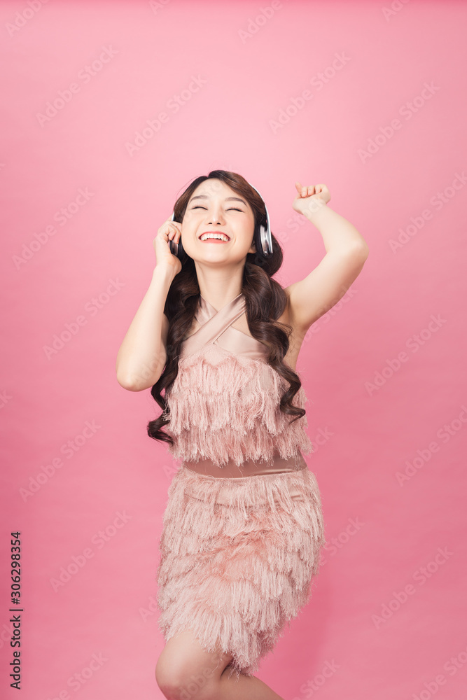 Happy joyful asian woman in headphones listening to music and dancing isolated over pink background