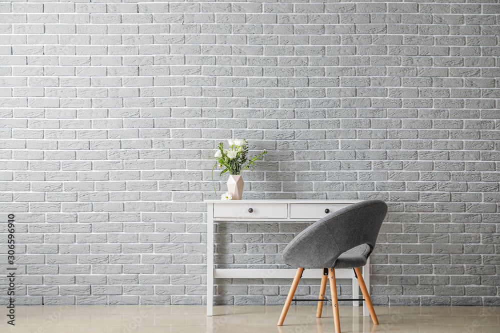 Fresh flowers in vase on table and armchair near brick wall