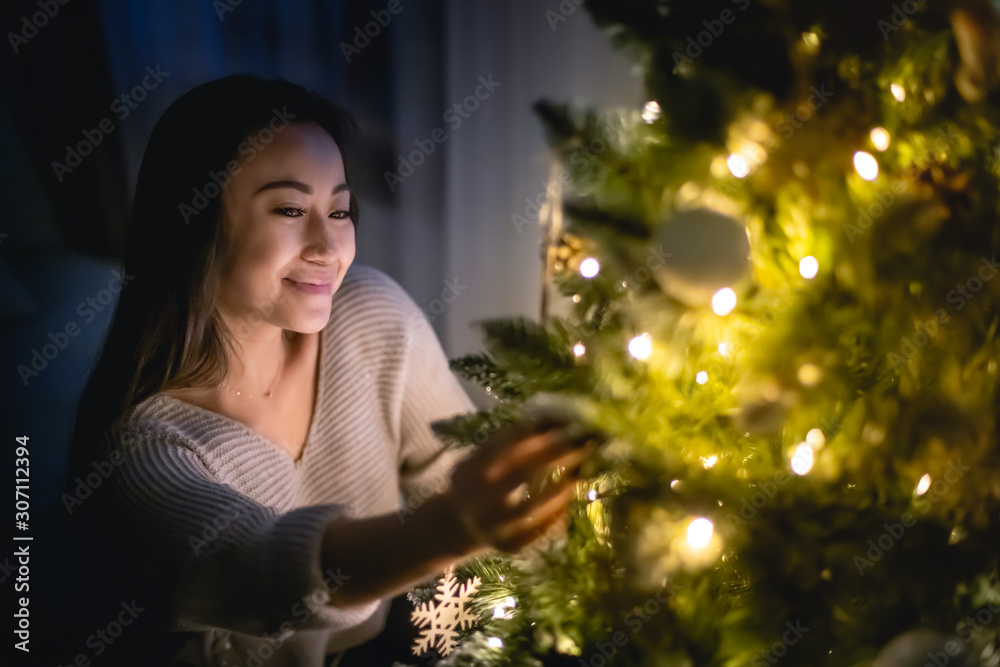 Beautiful Asian woman decorating Christmas tree in home at evening