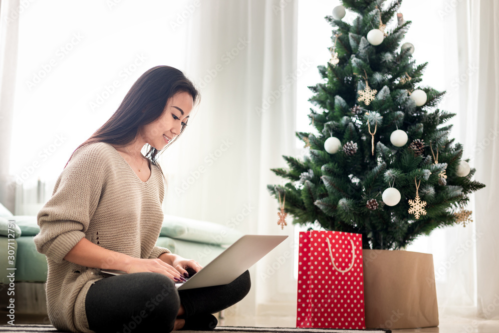 Happy asian girl shopping online with laptop sitting at decorated christmas tree background