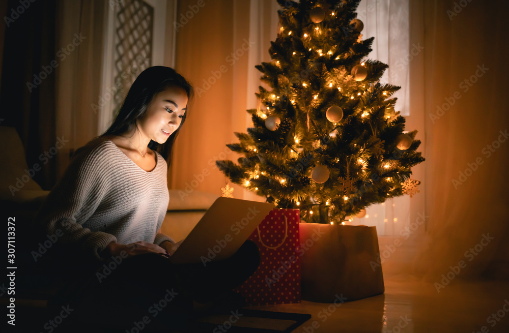 Happy asian girl shopping online with laptop sitting near decorated christmas tree background at eve