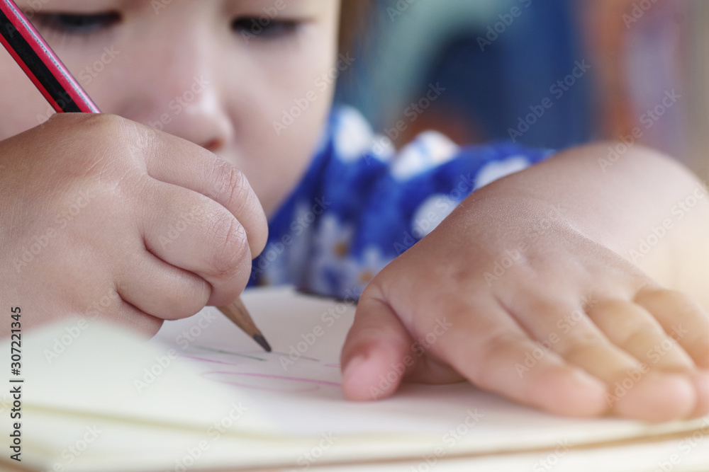 Close-up little girl child’s hand holding pencil drawing on notebook white paper, homeschool educati