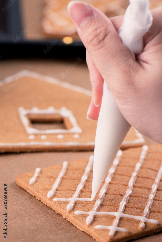 Woman is decorating gingerbread cookies house with white frosting icing cream topping on wooden tabl