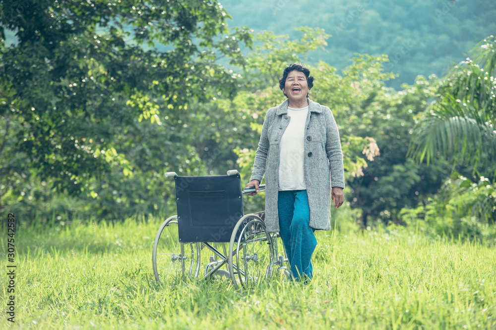 老年女性患者站在公园的轮椅旁微笑着。