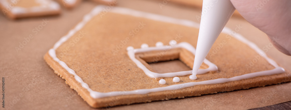 Woman is decorating gingerbread cookies house with white frosting icing cream topping on wooden tabl