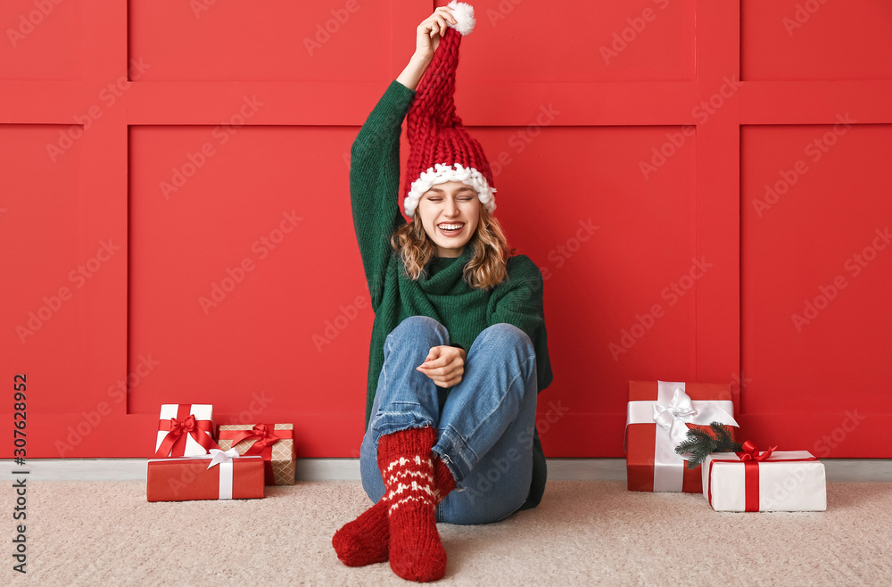 Happy young woman in winter clothes and with Christmas gifts sitting near color wall