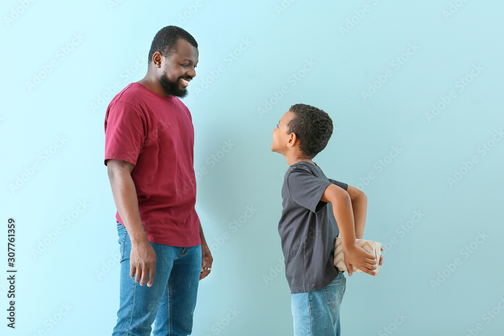 Little African-American boy hiding gift from his father behind back against color background