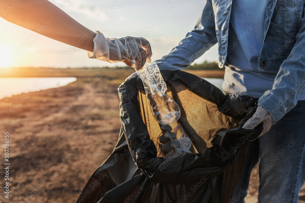 people volunteer keeping garbage plastic bottle into black bag at park near river in sunset