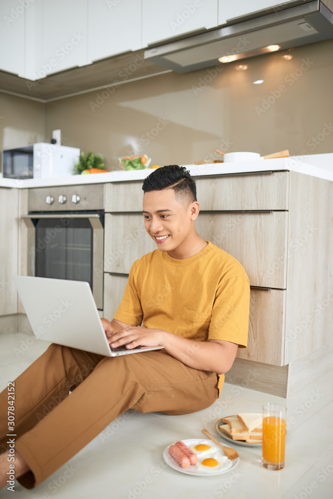 attentive asian man using laptop while sitting on floor in kitchen