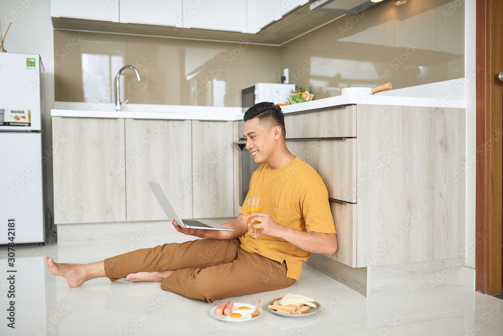 Young business man is smiling, eating breakfast and working on laptop in bright kitchen.